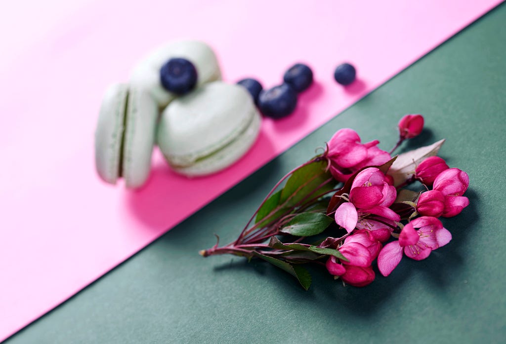 Image of green macarons with blueberries on a pink background, alongside a small bouquet of vibrant pink flowers on a green background. The contrasting colors create a visually appealing and elegant composition.