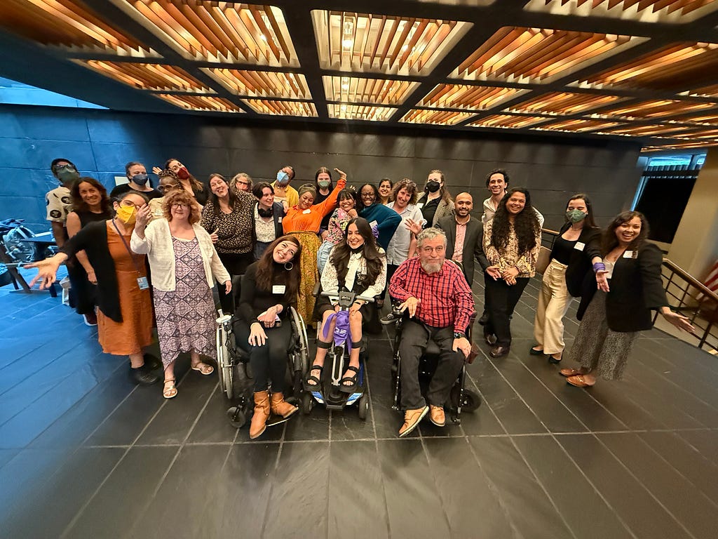 A group of people smile for the camera, standing in a large room with wooden ceiling panels. The picture features some of the attendees of WEI’s Legal Capacity Workshop.