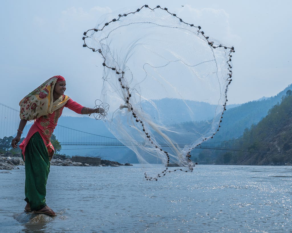 A woman tosses a fishing net in the air as she stands on a stone in the middle of a river.