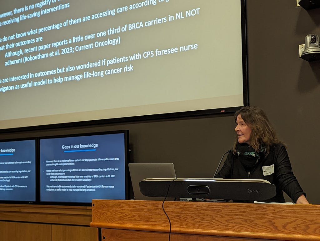 In a lecture hall, Dr. Holly Etchegary gives a talk at SHARE Summit 2023. Dr. Etchegary is standing at a podium facing the audience. Behind Dr. Etchegary are multiple screens displaying a PowerPoint slideshow.
