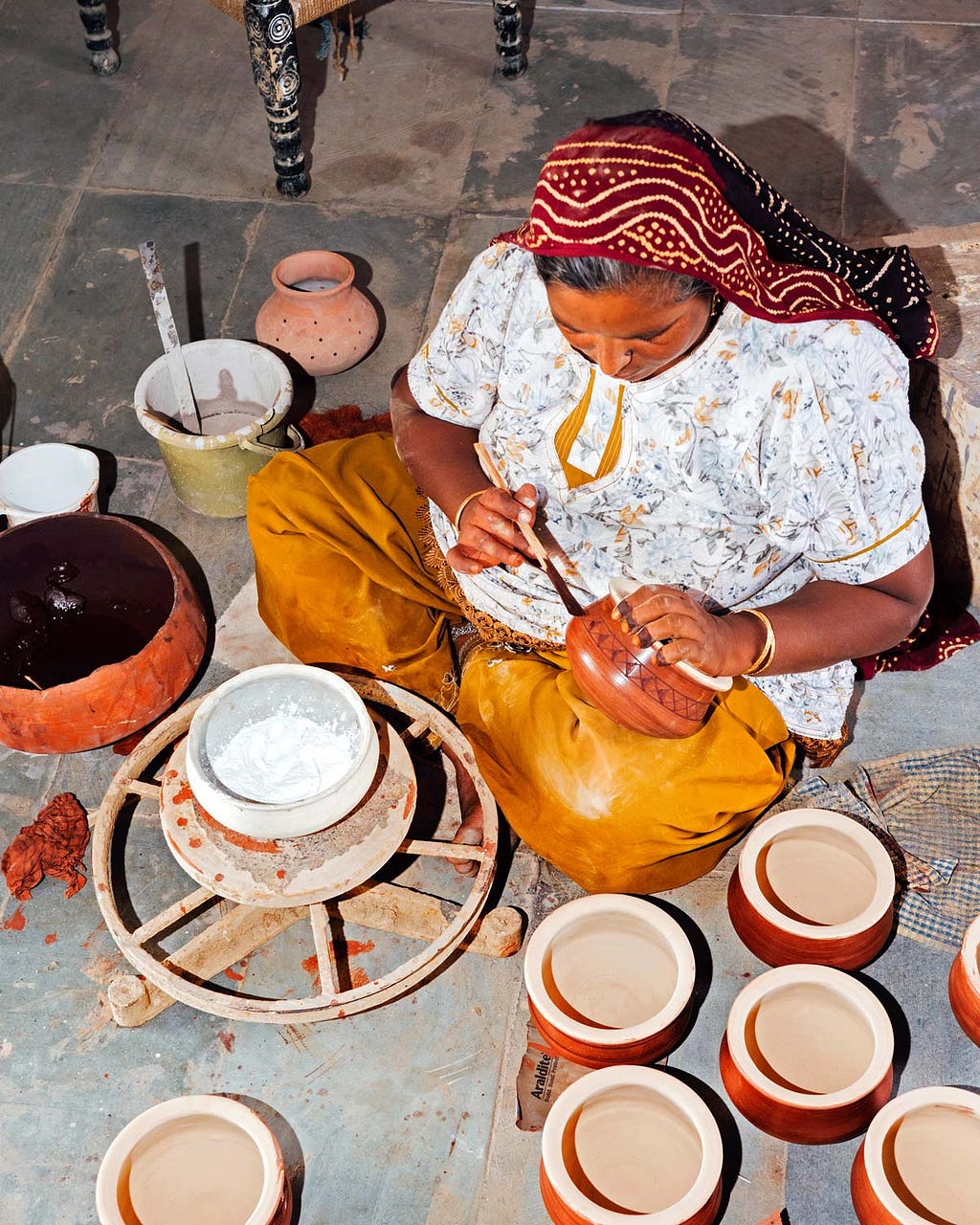 A woman sits cross-legged and paints finished pottery.
