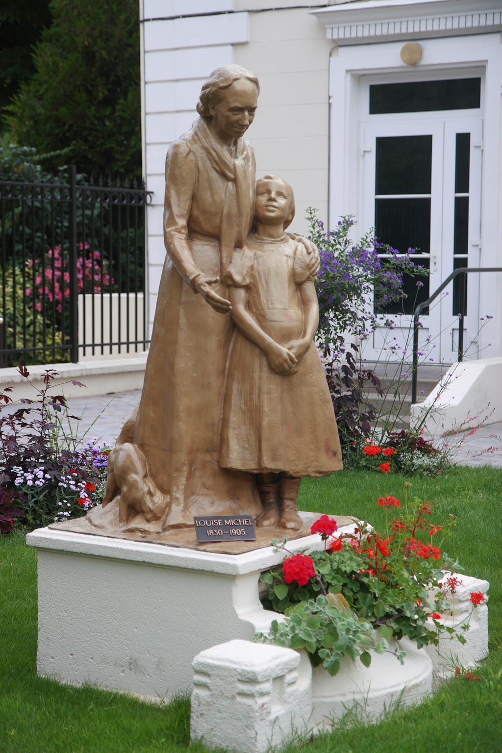In the bronze sculpture, an older Louise Michel stands with a hand on the shoulder of a young girl, holding her close, while making a gesture of encouragement. The girl seems to look up at the older woman with admiration, and on the base of the statue, a cat rubs lovingly up against Louise Michel.