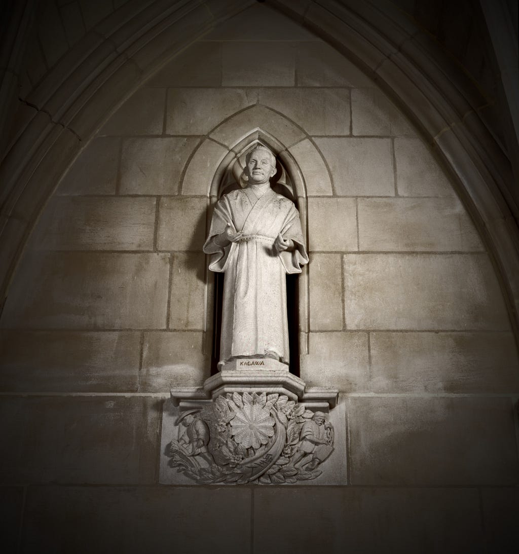 A carved limestone figure of Japanese pacifist Toyohiko Kagawa, originally sculpted by artist Frederick Hart. The figure wears an expression of understanding, his hands held forward, and partially open, as if in a gesture of giving. The figure is positioned in an arched niche in the center of the stone wall of the cathedral.