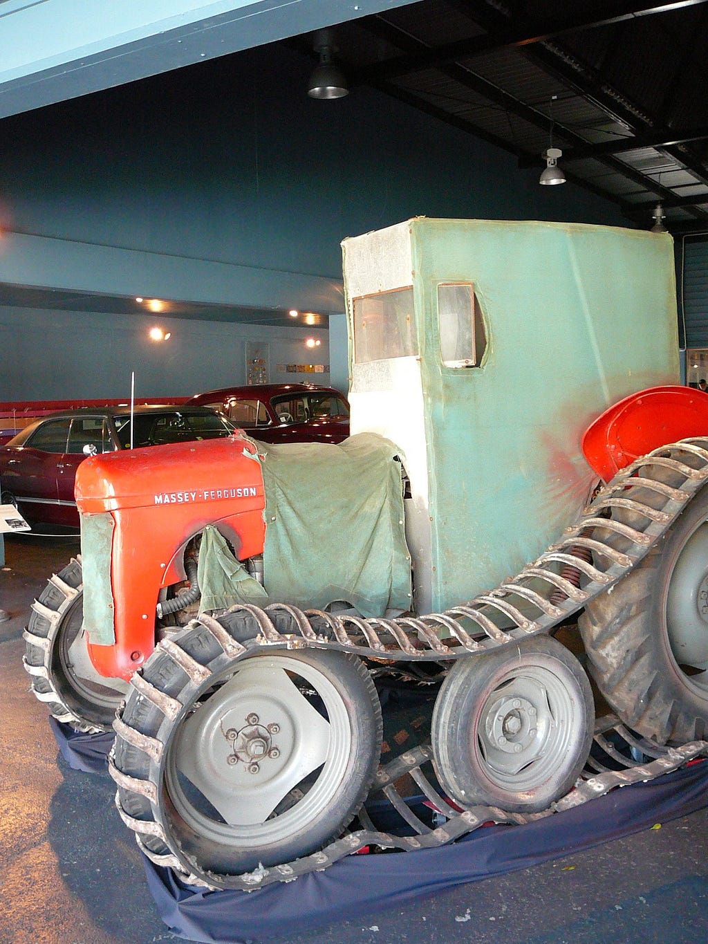 A red Ferguson farm tractor converted for use in Antarctica. There are metal tracks over the wheels, green canvas cover over the radiator grills and vents and a green canvas box built for the driver to sit in to protect them from the cold.