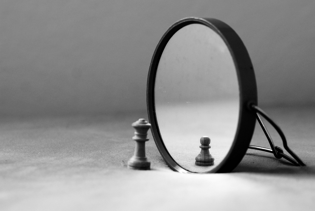 A black and white image of a knight chess piece in front of a small mirror, the reflection shows a pawn.