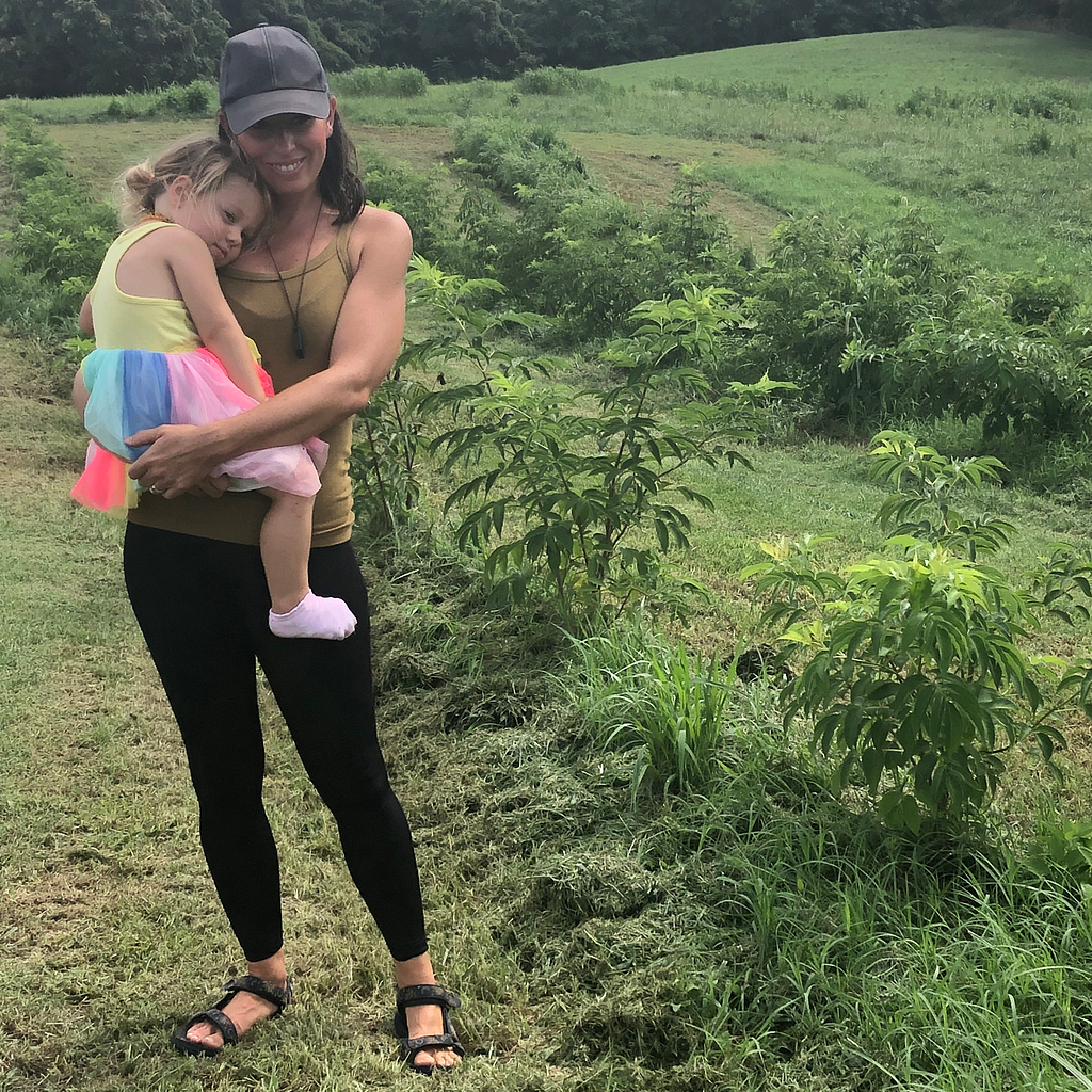 Kate, of Mama Bear’s Elderberries, stands to the left of the picture, holding her young daughter. They stand in front of rows of young elderberry trees.