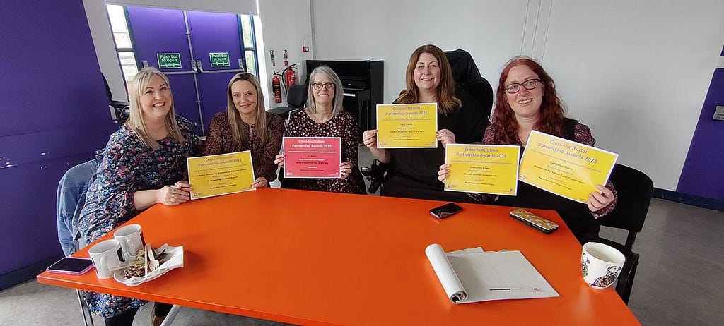 An image of Kim Crossley, Emma Taylor, Jo Huett, Faith Castle and Catherine Bates sat at a red rectangular table holding their local level Partnership Awards certificates.