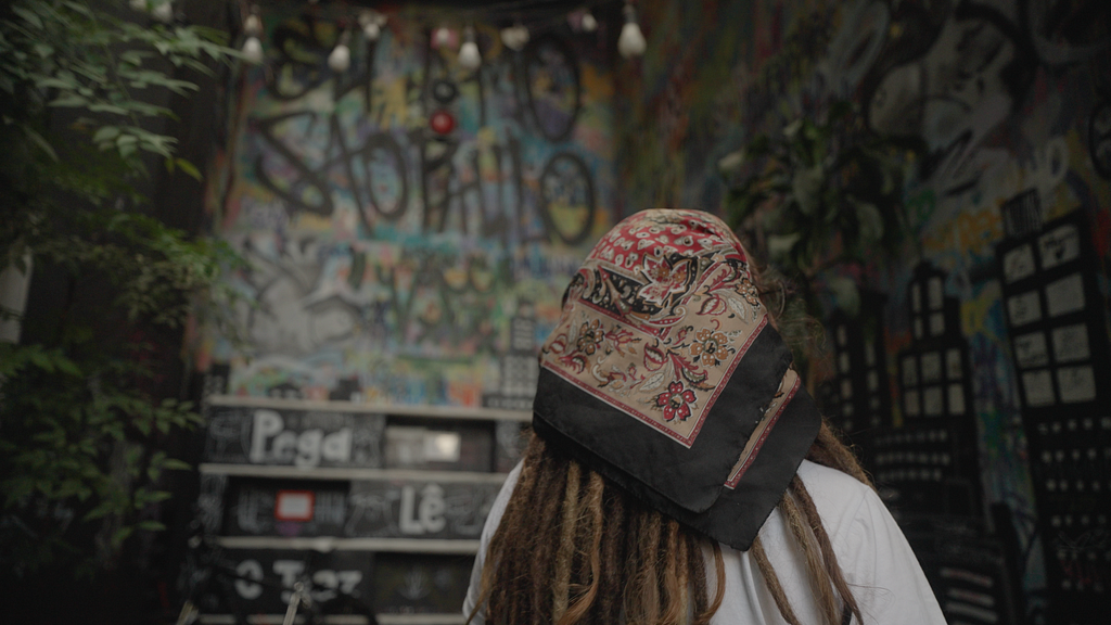 Foto de uma mulher de cabelos com dreads e lenço na cabeça. Ela está de costas para a câmera. Em frente a ela, está uma parede cheia de grafites.