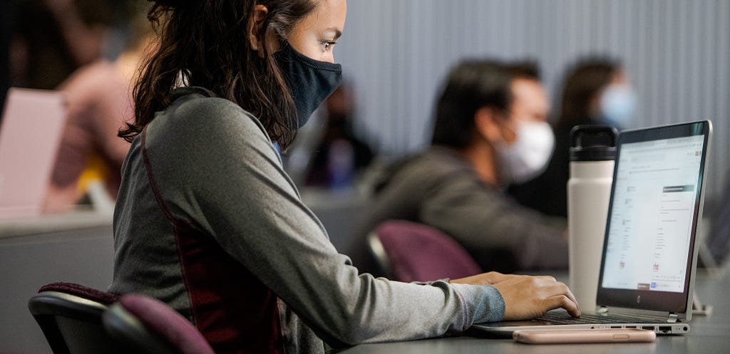 A woman wears a face mask while typing on her laptop. She is surrounded by other masked students in a lecture hall.