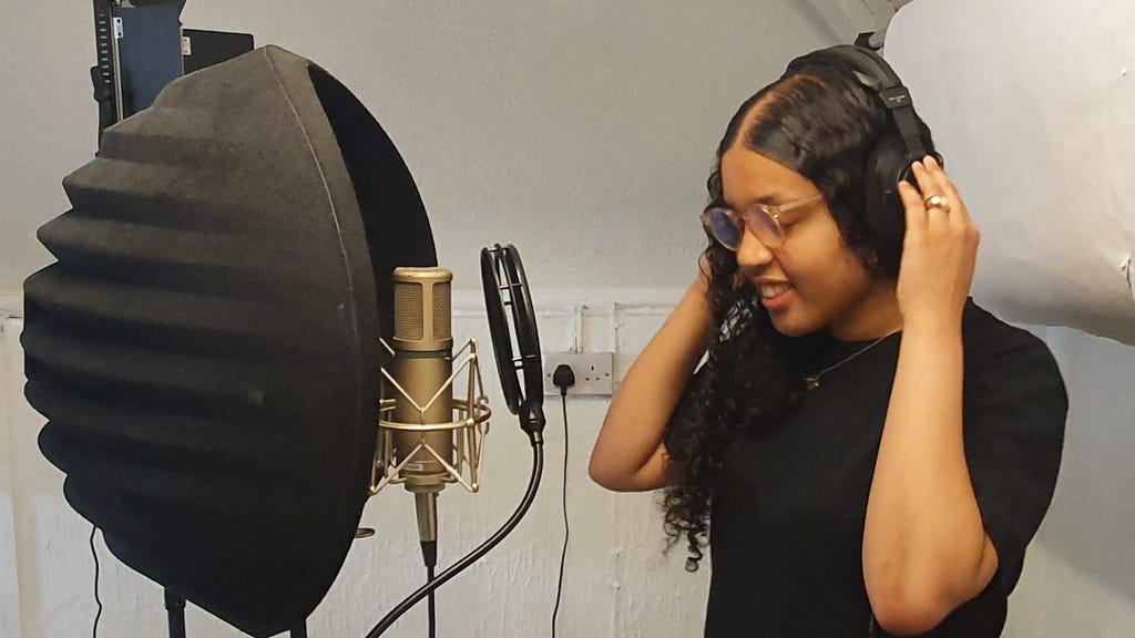 Malizah, standing next to recording kit, she is a oung Black woman with long black haid, and glasses. She is holding her headphones at each ear.