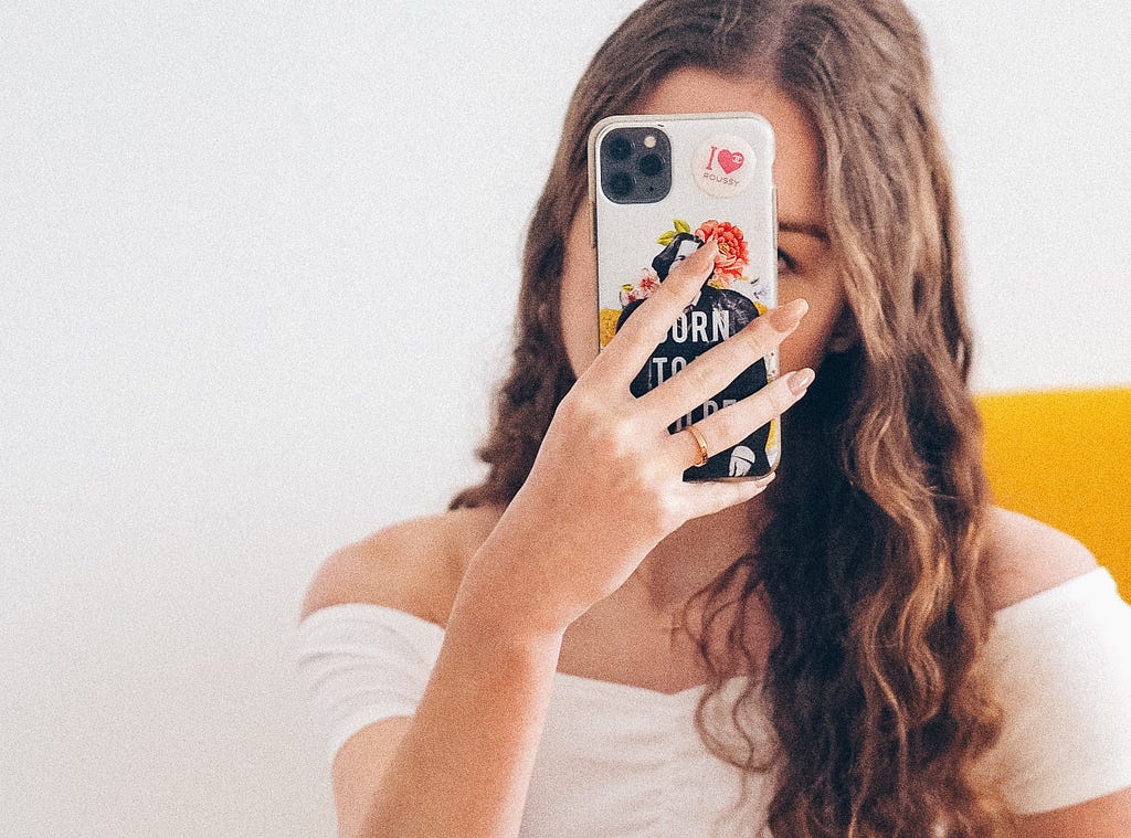White woman with long brown hair, wearing a white shirt and jeans, takes a selfie in the mirror with the camera covering her face.