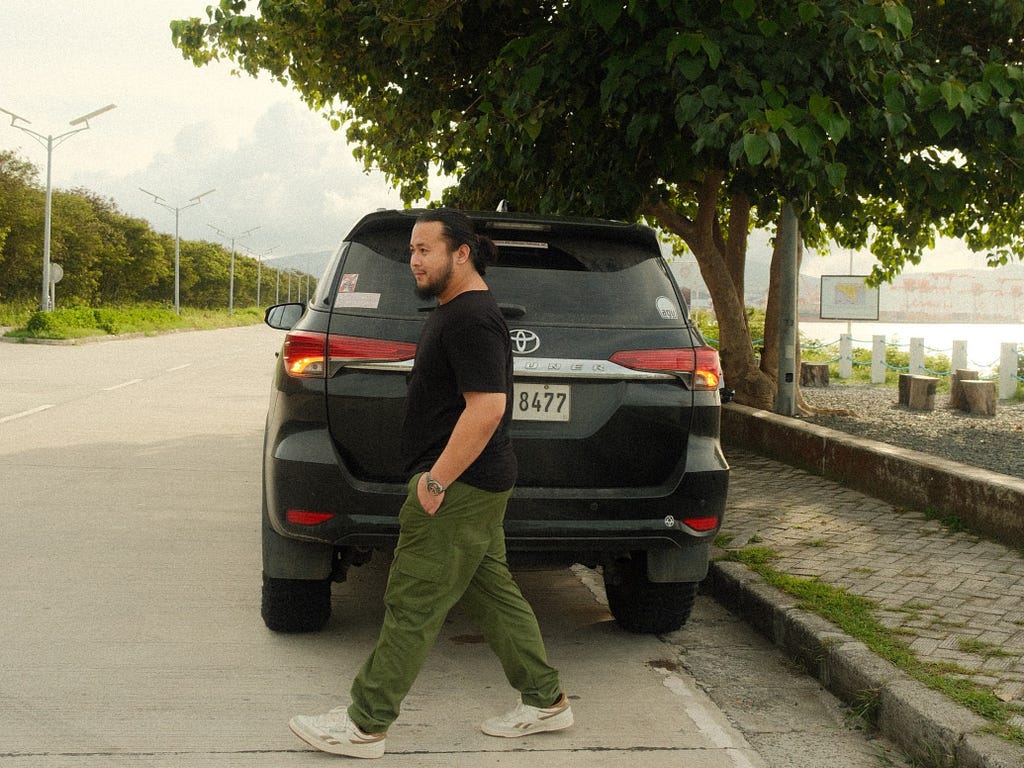 A man in a black shirt and cargo pants walking behind a car.