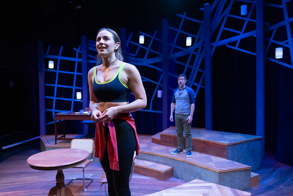 A woman in workout clothes stands on the set of THE WANDERERS speaking out, while a man wearing a baseball tee looks on from another part of the set.