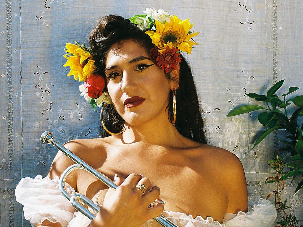 Headshot photograph of Latinx musician La Doña, wearing bright yellow and red flowers at the crown of her head and holding a trumpet against her chest, looking directly into the lens