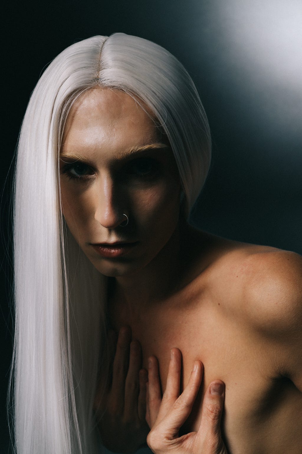 Buffy, close to the camera, intently stares forward wearing long white hair, her shoulders exposed with her hands covering her chest.
