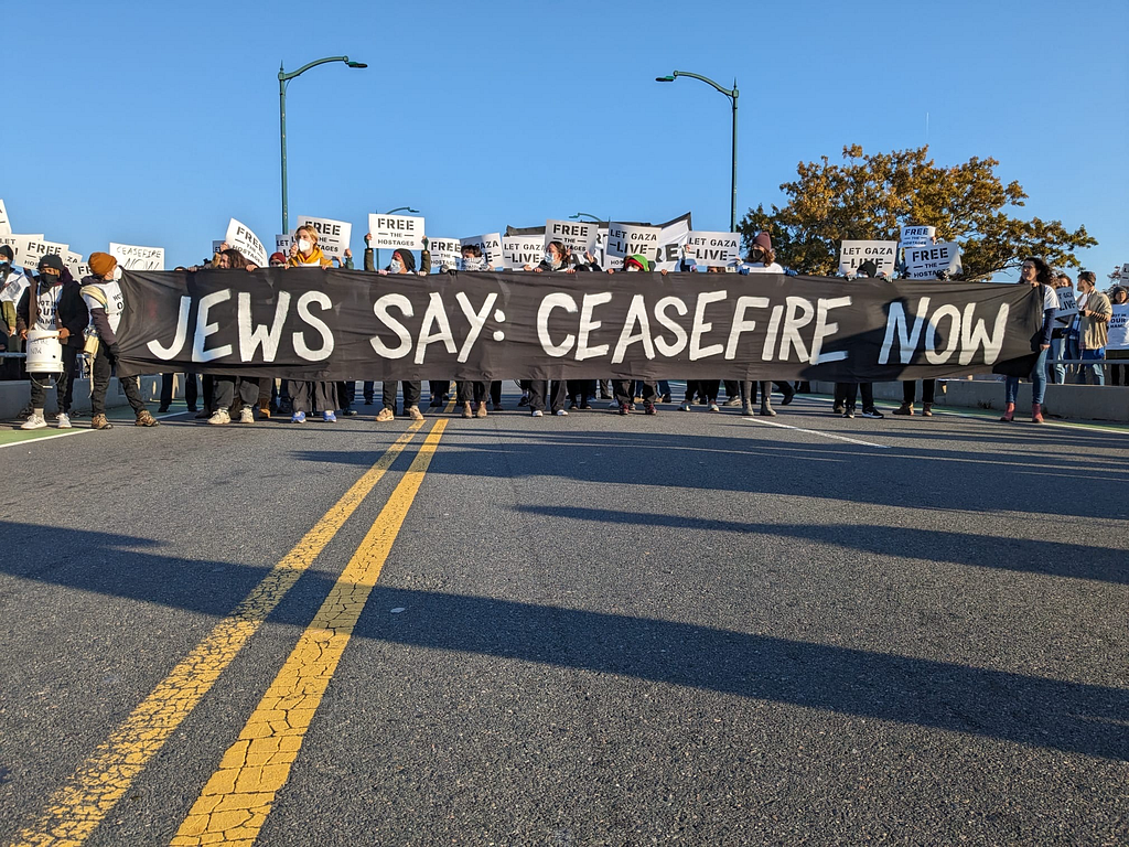 A road is completely blocked by a line of protesters. They’re holding a banner that says “JEWS SAY: CEASEFIRE NOW”