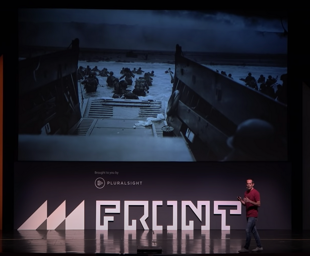 Wade Shearer on stage with a picture of soldiers running out of a boat towards the beach on D-Day in the background.