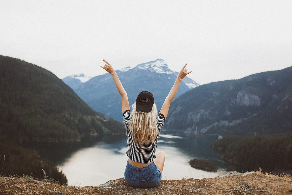 woman sitting on cliff raising both hands, self-confident and looking at mountains and a lake