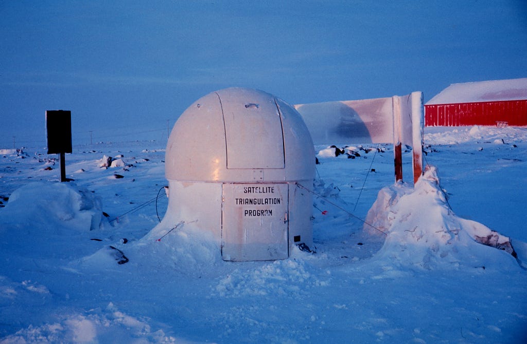 Obligatory blog header photo. This one is a snowy, rocky landscape. In the center is white domed NOAA station with 2 hatches. The bottom hatch says SATELLITE TRIANGULATION PROGRAM. There’s a red shed in the background.