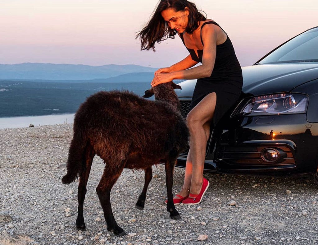 A woman in a black dress petting a black sheep on a rocky roadside at sunset, next to a black car — an unexpected, playful moment in an unusual setting.