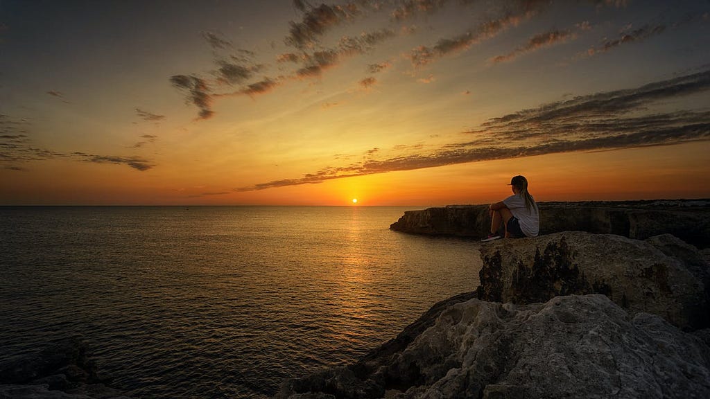 a woman sitting on the rocks besides sea and watching sunset and the skyline