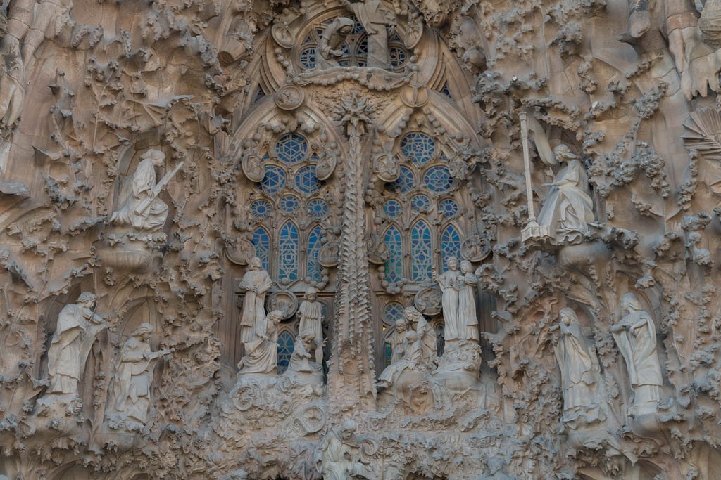 Sculptures of people surround blue tinted windows on the outside of the Sagrada Família.