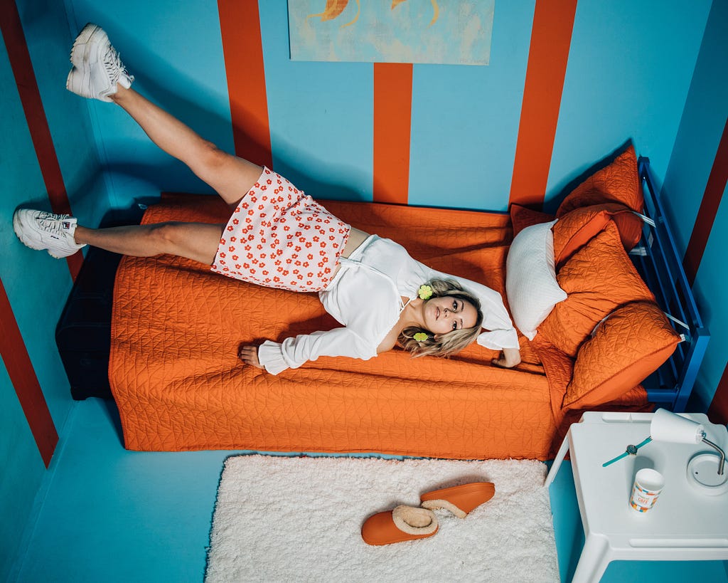 Girl awkwardly lying on orange bed with her feet on the blue and orange striped wall in a tiny room
