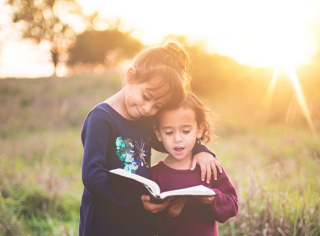 Two girls stand in a sunlit field with a book, one reading from the book and one smiling
