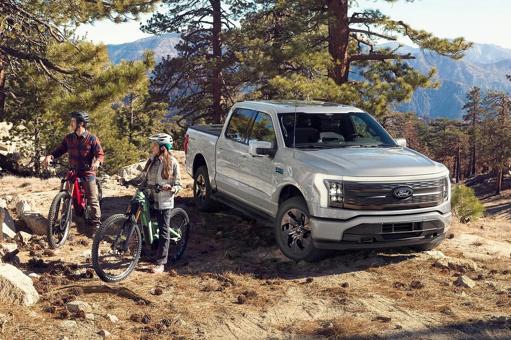 From promotional materials, two mountain bikers standing next to their bikes in a scenic forested area, beside a silver Ford F-150 Lightning electric truck parked on rugged terrain with mountains in the background