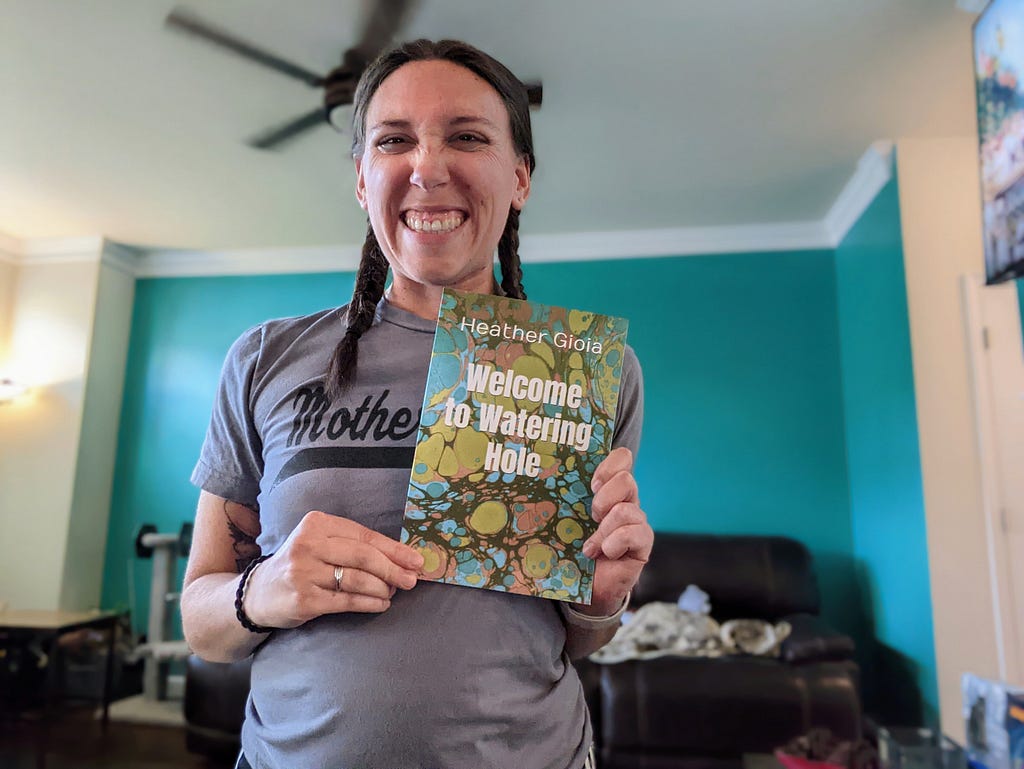 Heather Gioia, author of “Welcome to Watering Hole”, is holding a print copy of the book with a big smile. Heather is in a gray t-shirt with braids in her hair. She is standing with green and white walls in the background.