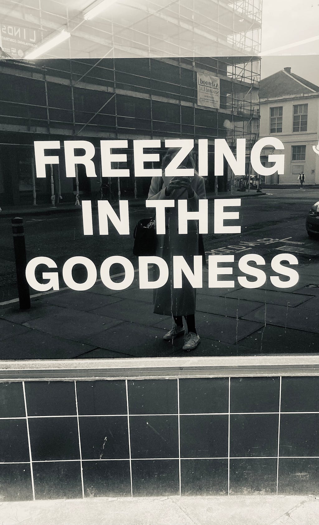 A black and white photograph of my reflection in a window, which says ‘freezing in the goodness’. It was a particularly cold day in Edinburgh.