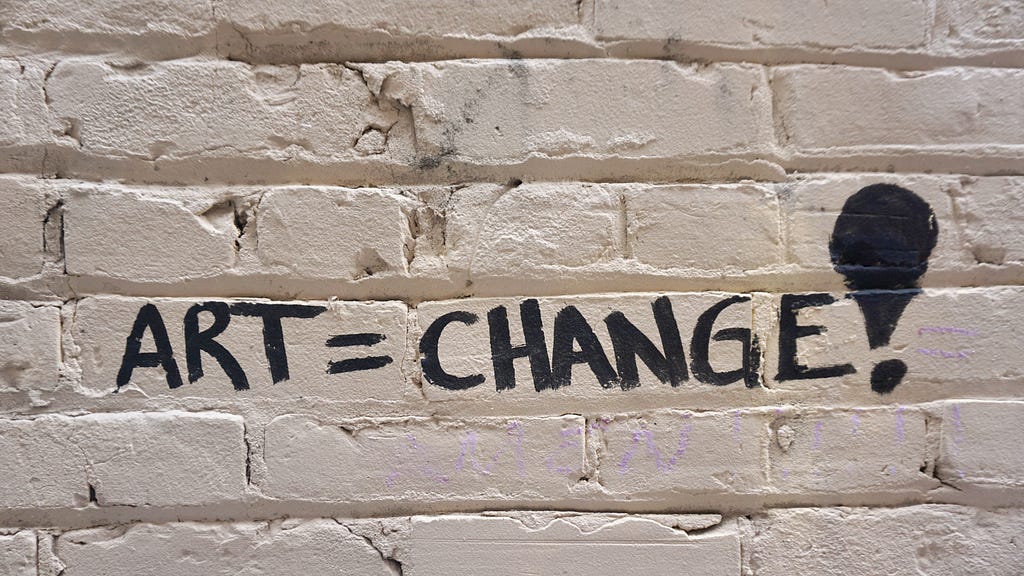The words “ART = CHANGE!” are hand-written on a white brick wall