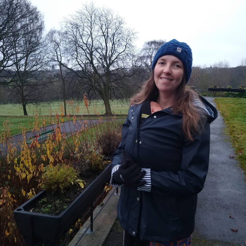 The author is pictured. She has long brown hair, is white and is wearing a blue beanie hat and coat. Peeping out from her coat collar you can see the head of a small baby in a wrap. They are in a park, in the background are trees and shrubs.
