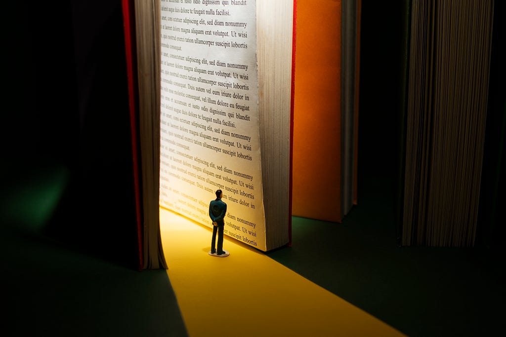 Um pequeno boneco olha para a página de um livro, que está “de pé‘. De dentro do livro sai um feixe de luz, que ilumina o boneco e a página.
