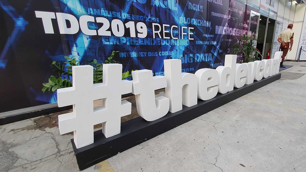 Imagem mostra painel escrito TDC2019 RECIFE e #thedevconf no chão com letras de madeira