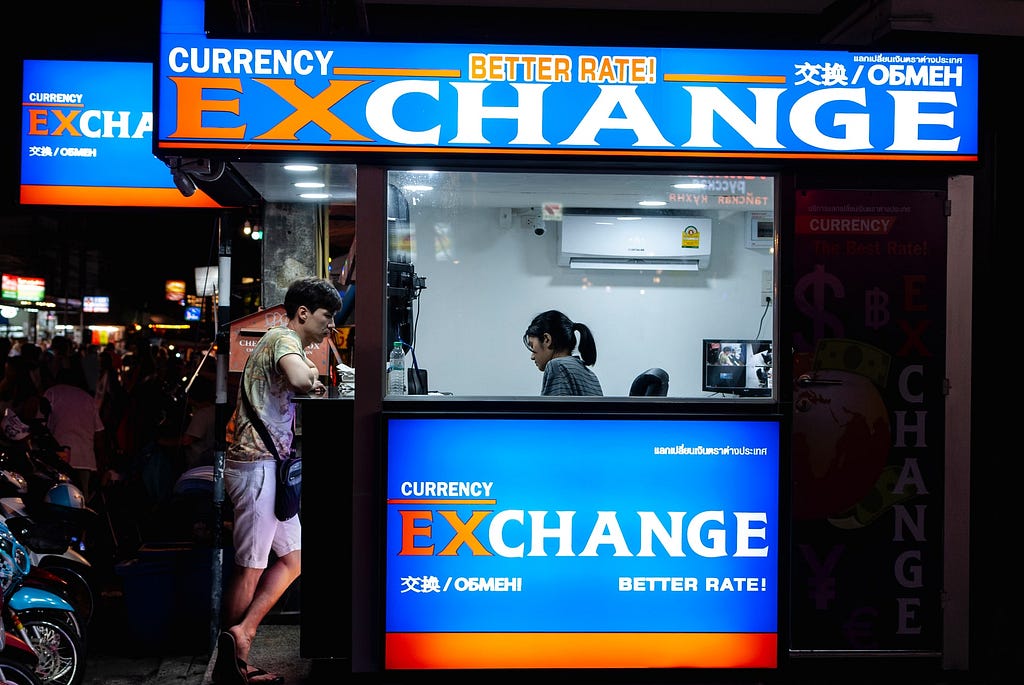 Man leaning on a currency exchange kiosk counter at night while a woman inside helps him.