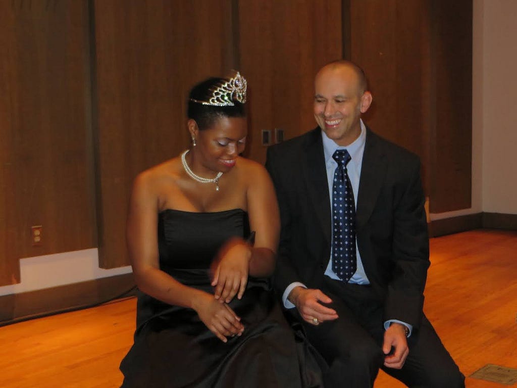 A happy couple in formal attire. A woman wearing a black gown, a pearl necklace, and an extravagant tiara. A man wearing a suit and tie. They are both smiling.