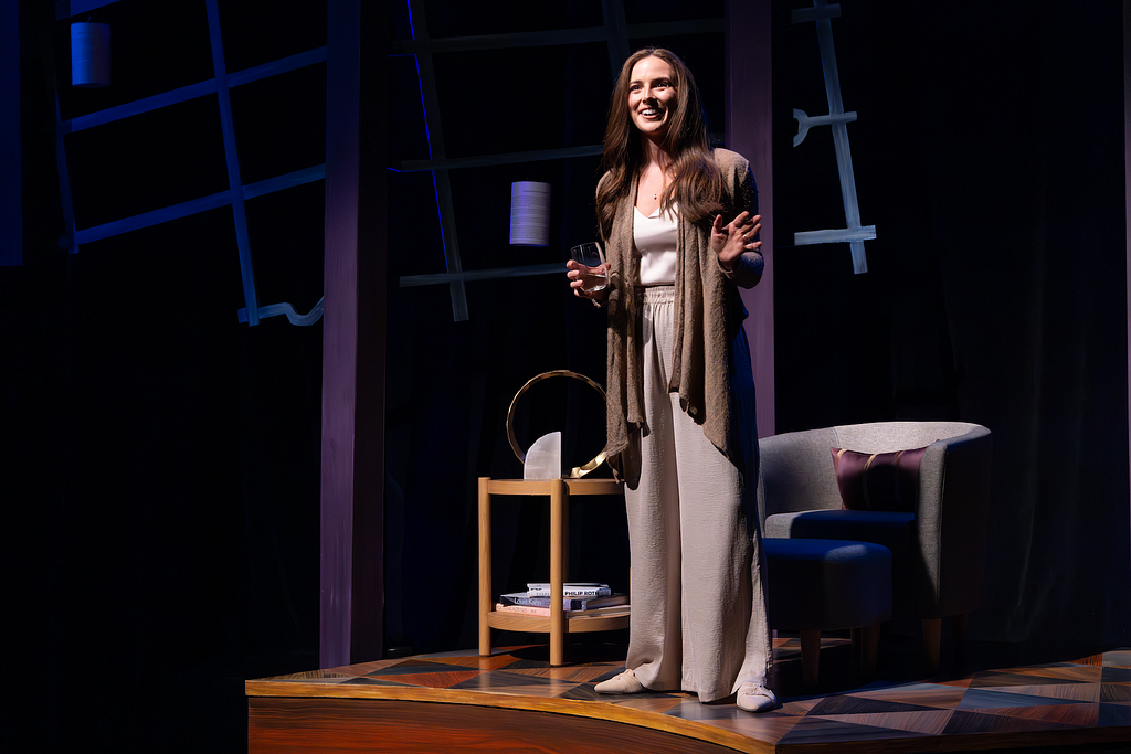 An actress stands onstage with a glass of wine, speaking and smiling. Her hair is down and she is wearing luxurious, flowy clothes.