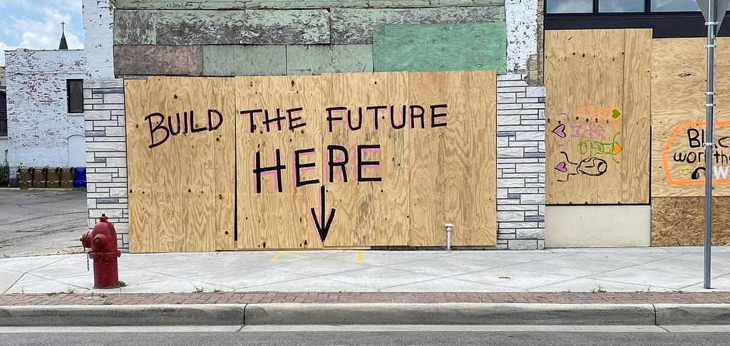 A boarded-up entrance has “Build the future here” painted on it, with an arrow pointing to the sidewalk below
