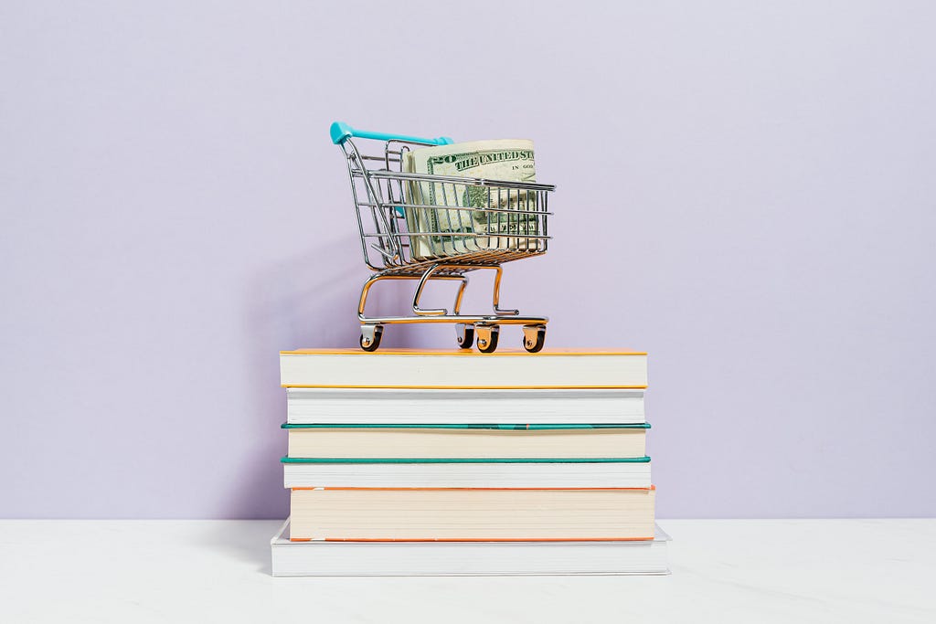 A toy shopping cart filled with US dollars sits on top of a stack of books.