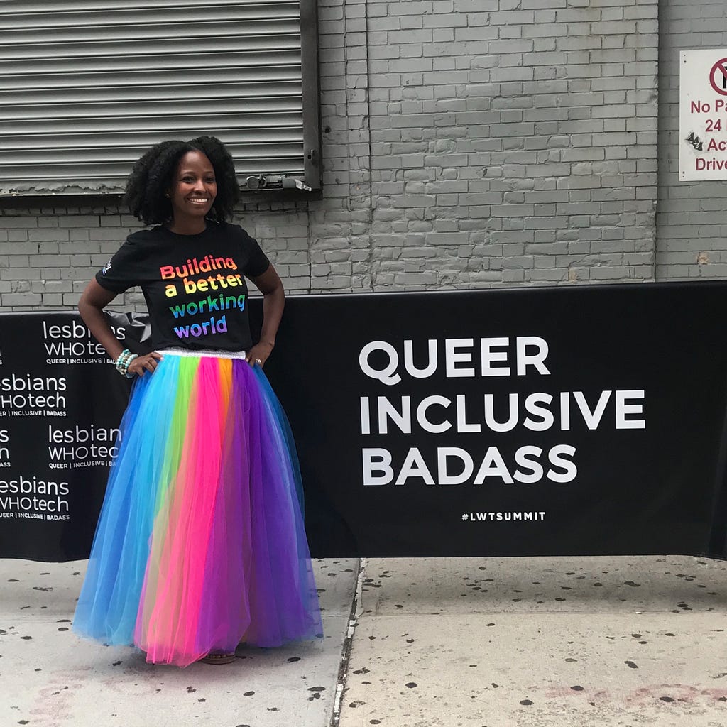 Summit attendee with a “Building a better working world” t-shirt, standing next to a sign that reads “Queer Inclusive Badass”