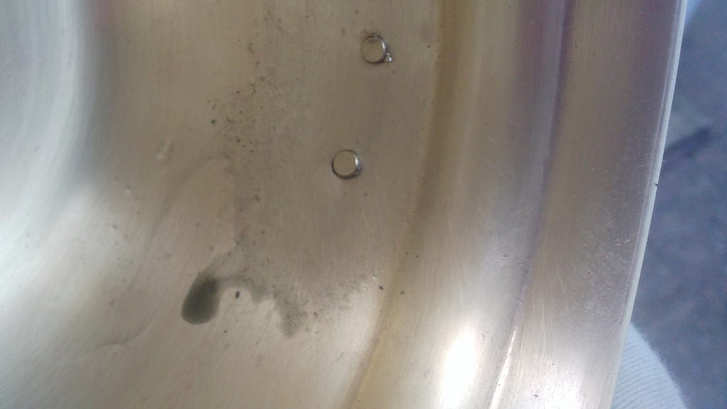Close up photo shows the inside of a brass pot where dirty lemon juice is seeping out of crevices around the rivets of the handle, having been gently loosened with a brass wire brush.