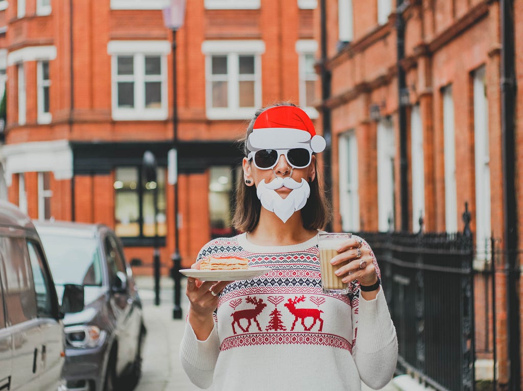 woman wearing a Christmas jumper and wearing a Santa mask, holding filled cup and festive pastry standing outside on a street