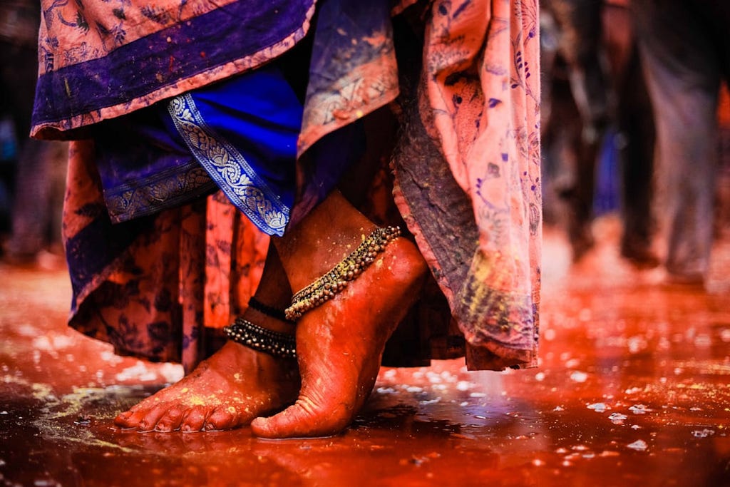 A cropped image of a woman’s feet standing in a shallow puddle with wide silver anklets and an overhanging sari.