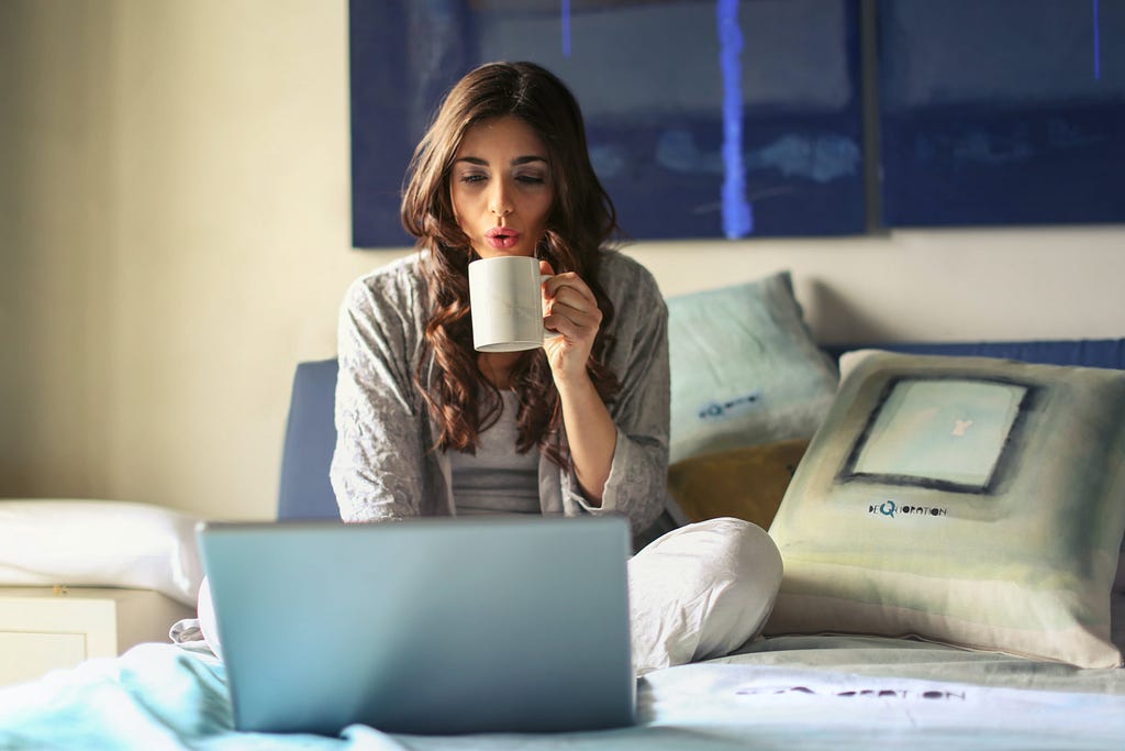 A woman in bed, sipping from a mug while typing on her laptop