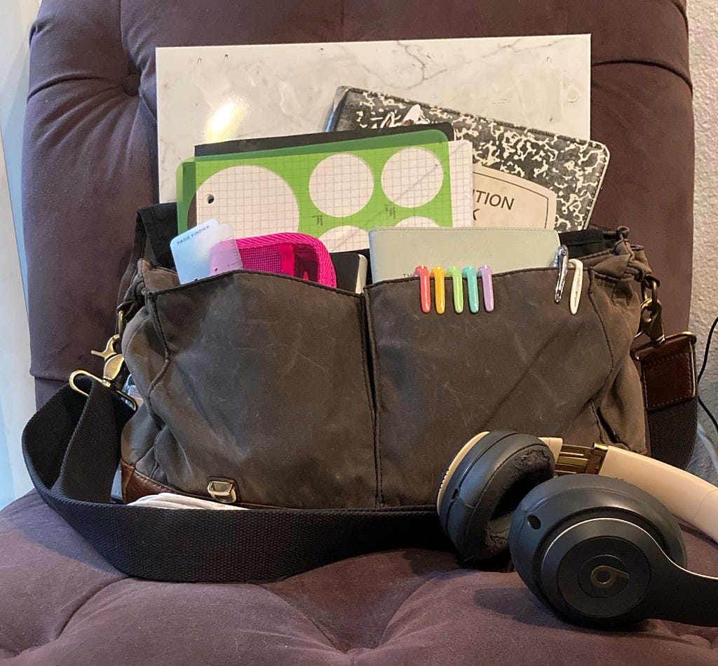 messenger bag filled with notebooks, pens, design stencils sits on a purple chair with a macbook charger and headphone