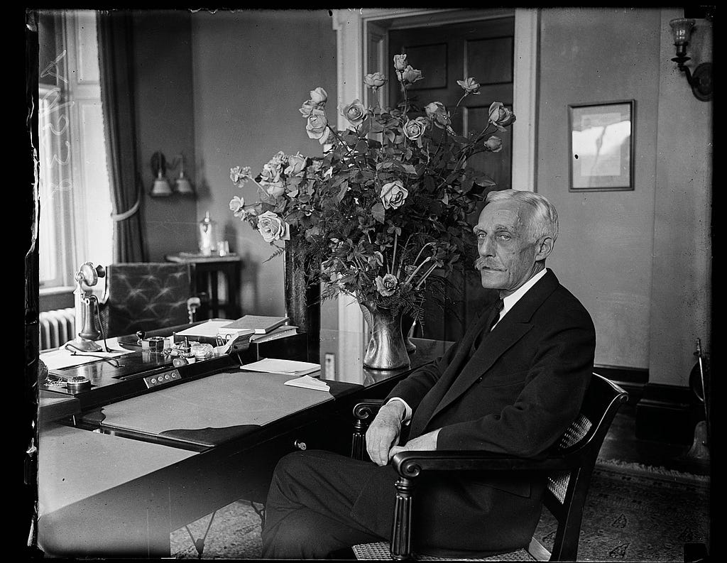 A b/w photo of Andrew Mellon sitting at a desk with a large floral arrangement behind him.