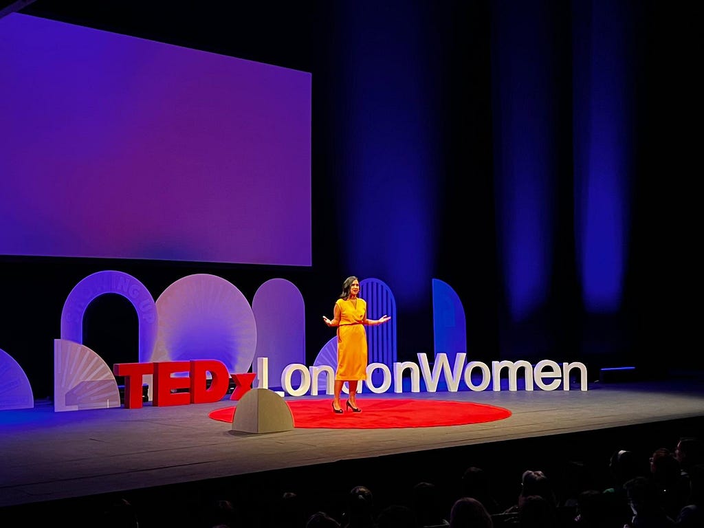 Anneka on stage wearing yellow dress, with arms opened out. Standing on big red circular carpet on a black stage. Behind are the letters ‘TEDxLondonWomen’ against purple-blue stage lights.