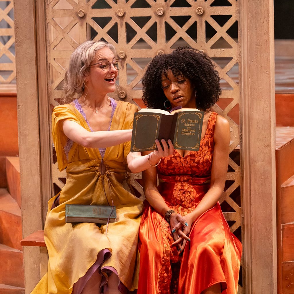 A woman in a yellow dress excitedly shows a book to a woman in an orange jumpsuit who reads sadly.