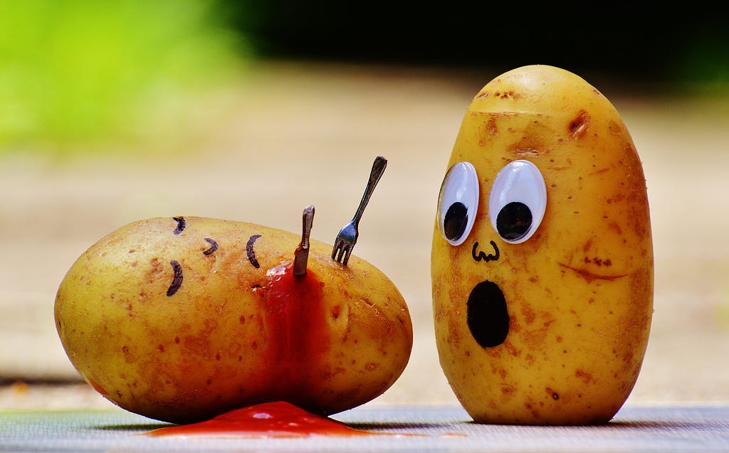 A potato with a drawn on face has been killed by a tiny knife and fork. It is bleeding out ketchup. Another potato looks on, shocked.